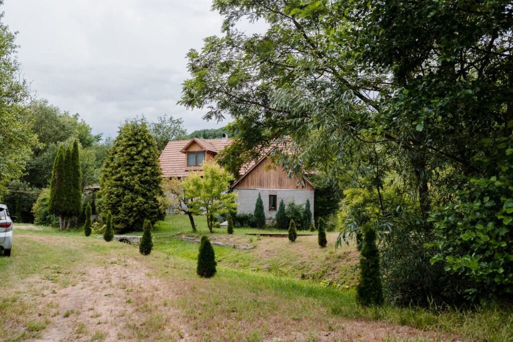 Vedere de pe drumul de acces către Cabana Ana. Se observă anexa nouă a cabanei, cu pereți albi și acoperiș de țiglă, și o parte din cabana principală, situate de-o parte și de alta a unui pârâu mic. Peisajul este înconjurat de vegetație luxuriantă.