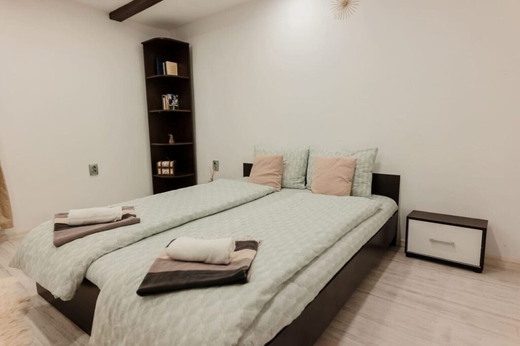 Imagine din Camera 1 de la parter, cu patul dublu aranjat pentru două persoane, o bibliotecă de colț din lemn închis la culoare și o noptieră modernă. Decorul este simplu, minimalist, cu perne roz și prosoape aranjate cu grijă pe pat.