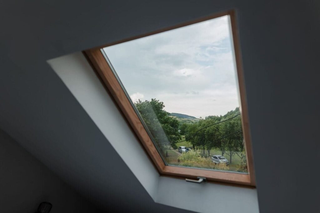O imagine din interiorul Camerei 3 de la mansardă, cu un geam Velux deschis. Se observă o priveliște superbă a curții cabanei, cu copaci, spațiu verde și mașini parcate, sub un cer acoperit de nori. Fereastra aduce lumină naturală în cameră și permite oaspeților să admire peisajul.