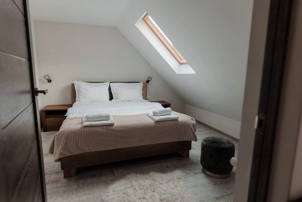 O vedere din holul de la etaj spre interiorul Camerei 3. Se observă un pat dublu, un luminator pe acoperiș și o noptieră modernă. Ușa de la intrare se deschide spre un spațiu curat, cu design minimalist, care invită la relaxare.