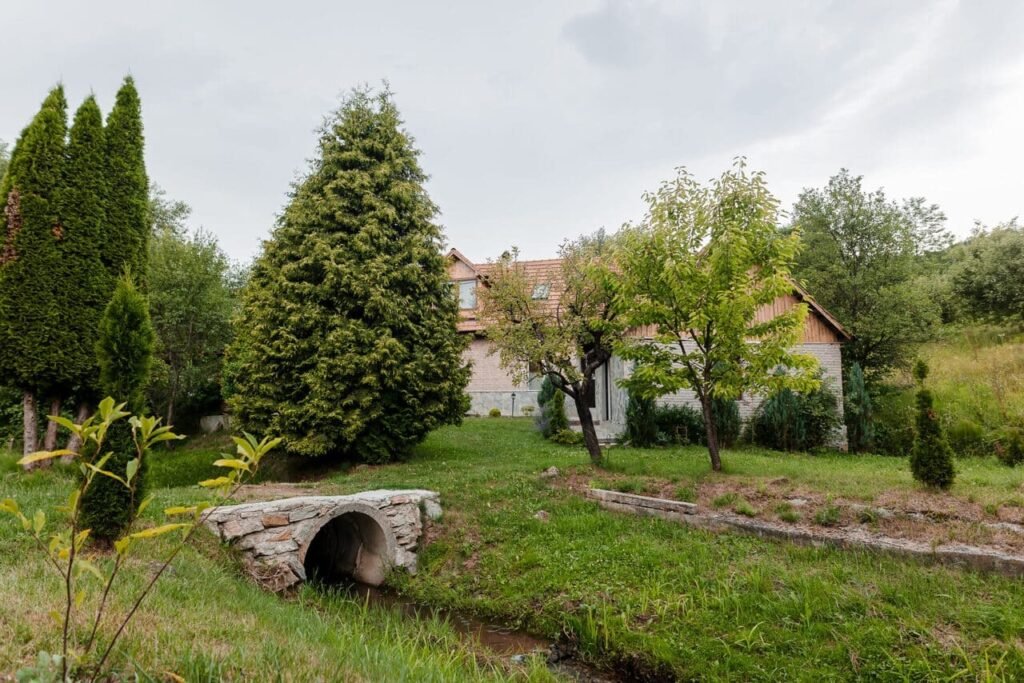 O imagine cu un pod de piatră rustică peste un pârâu mic, care asigură accesul spre anexa Cabanei Ana. Se observă anexa cu pereți din piatră, copaci verzi și un peisaj natural, care subliniază armonia și frumusețea proprietății.