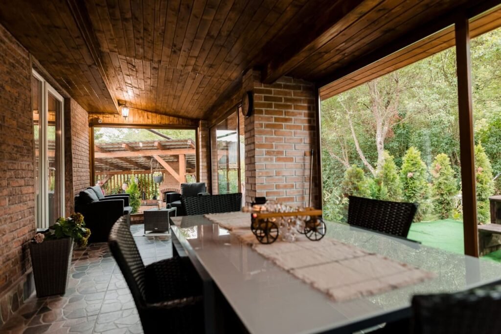 Imagine din interiorul terasei acoperite de la Cabana Ana, cu o masă mare, scaune elegante, și o canapea confortabilă. Se observă tavanul din lemn și pereții din cărămidă, cu o priveliște directă spre grădina verde și luxuriantă a proprietății.